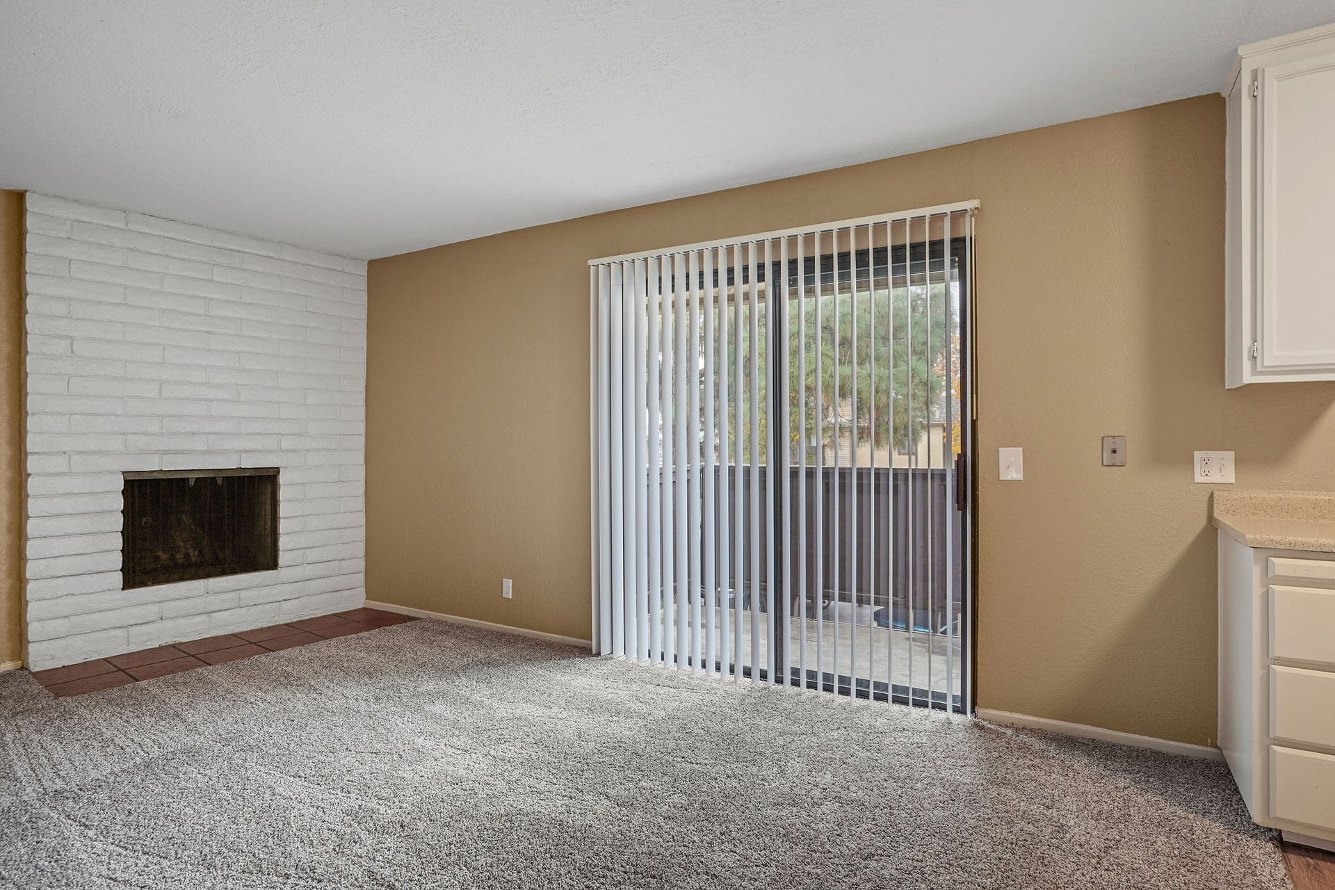 Empty living room with beige walls, white brick fireplace, carpet, and sliding glass door with vertical blinds