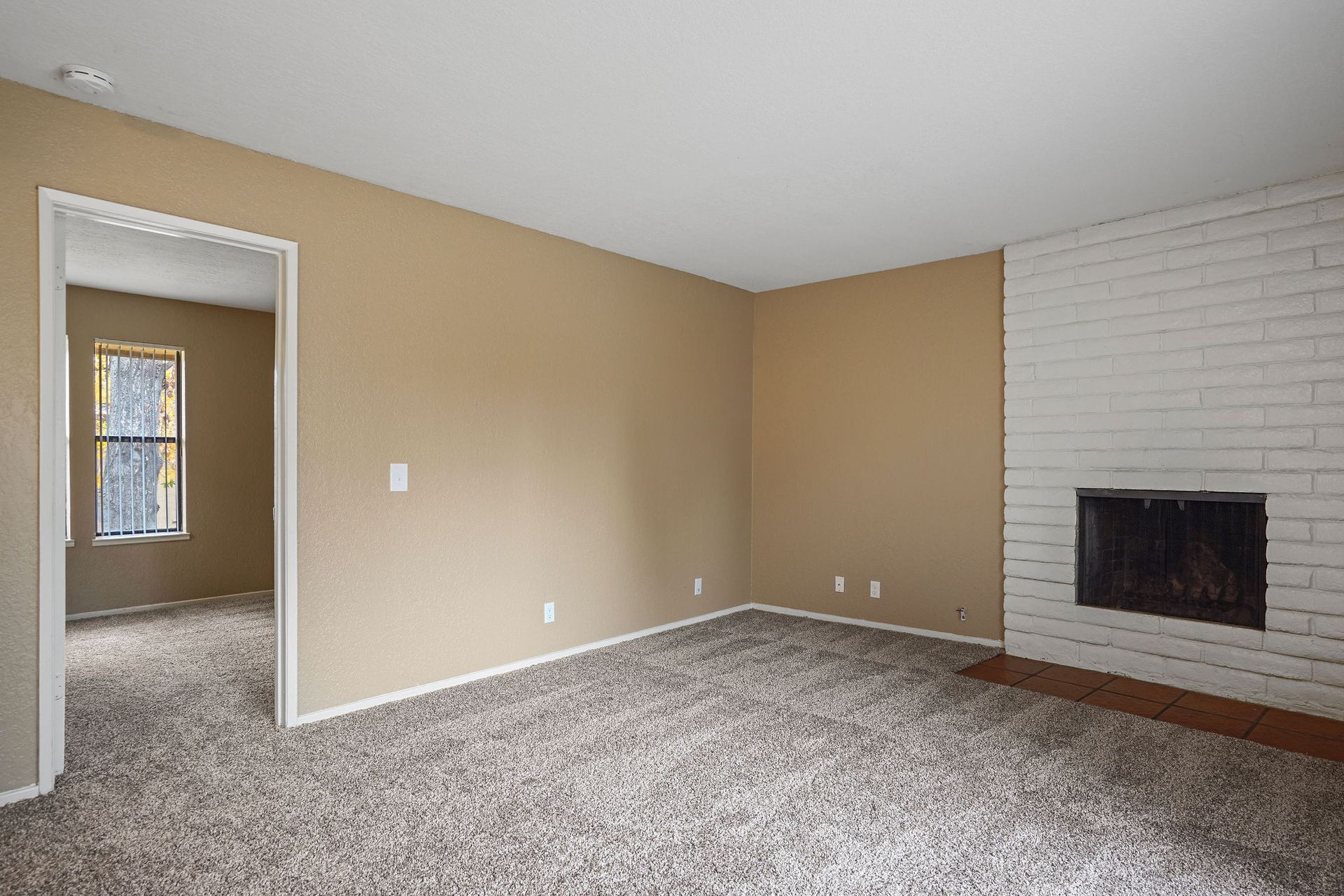 Empty beige living room with gray carpet, white brick fireplace, and doorway to another room