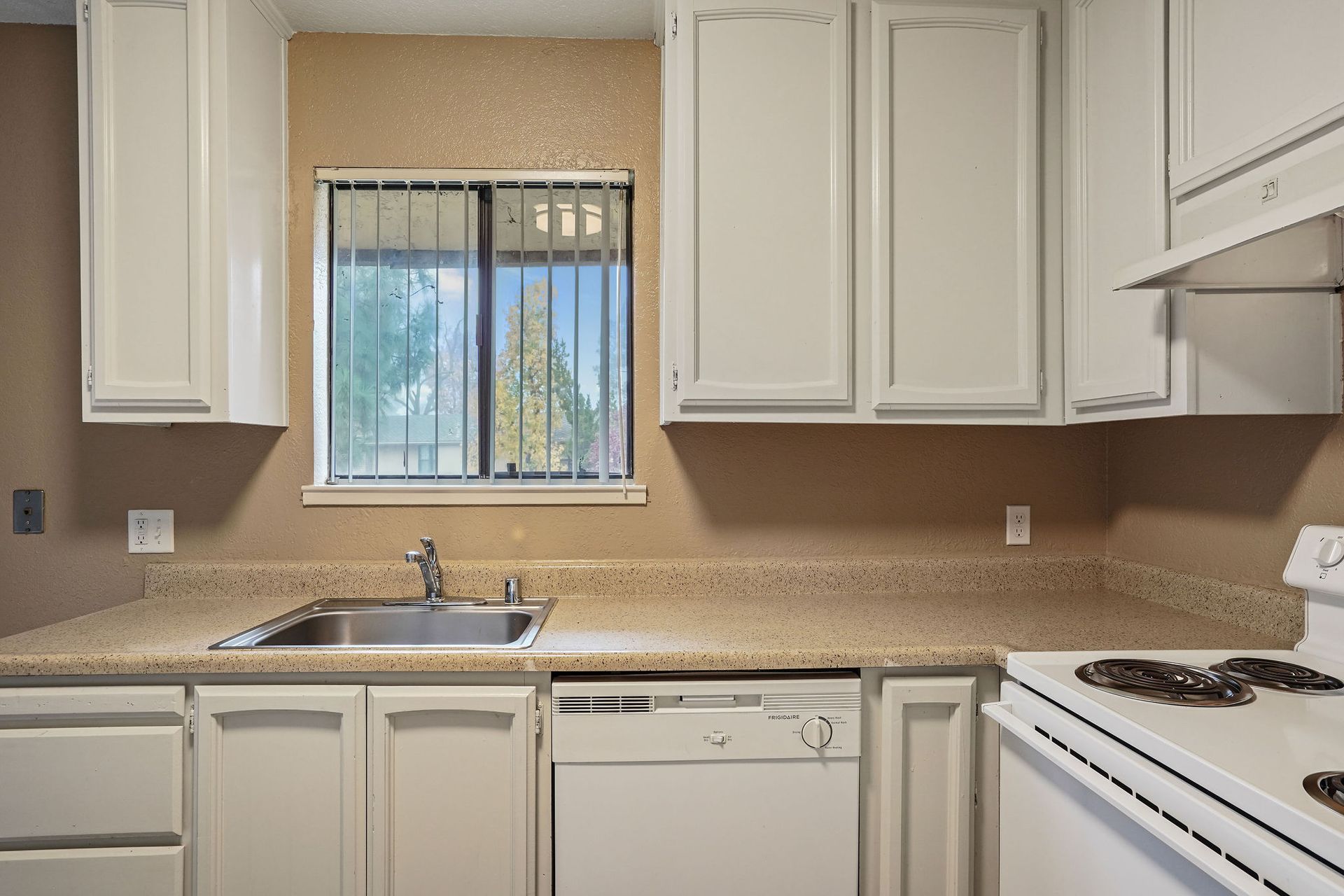 Empty kitchen with white cabinets, tan countertop, sink under window, dishwasher, and stove on the right