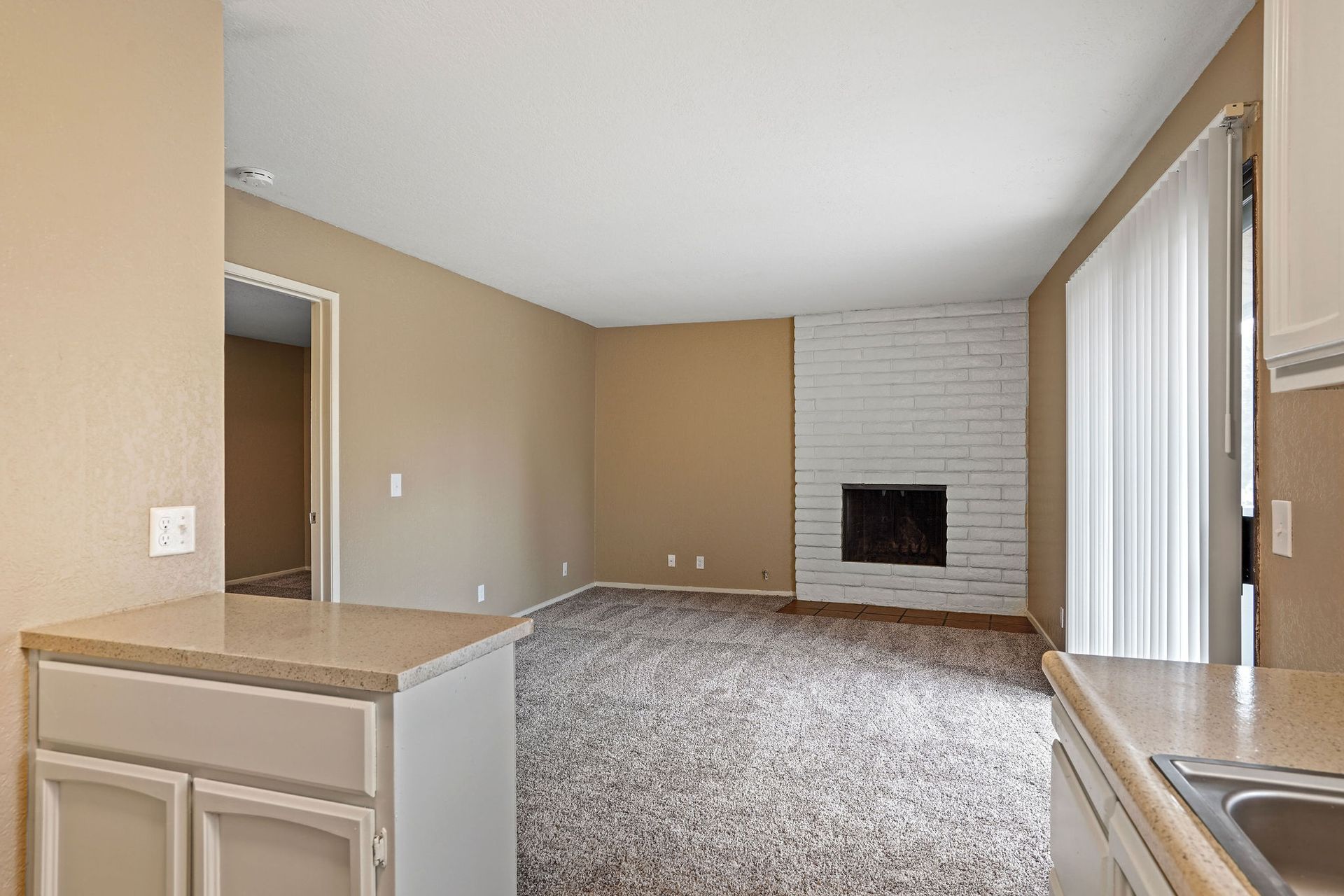 Empty living room with beige walls, carpet, and a white brick fireplace beside sliding glass doors.