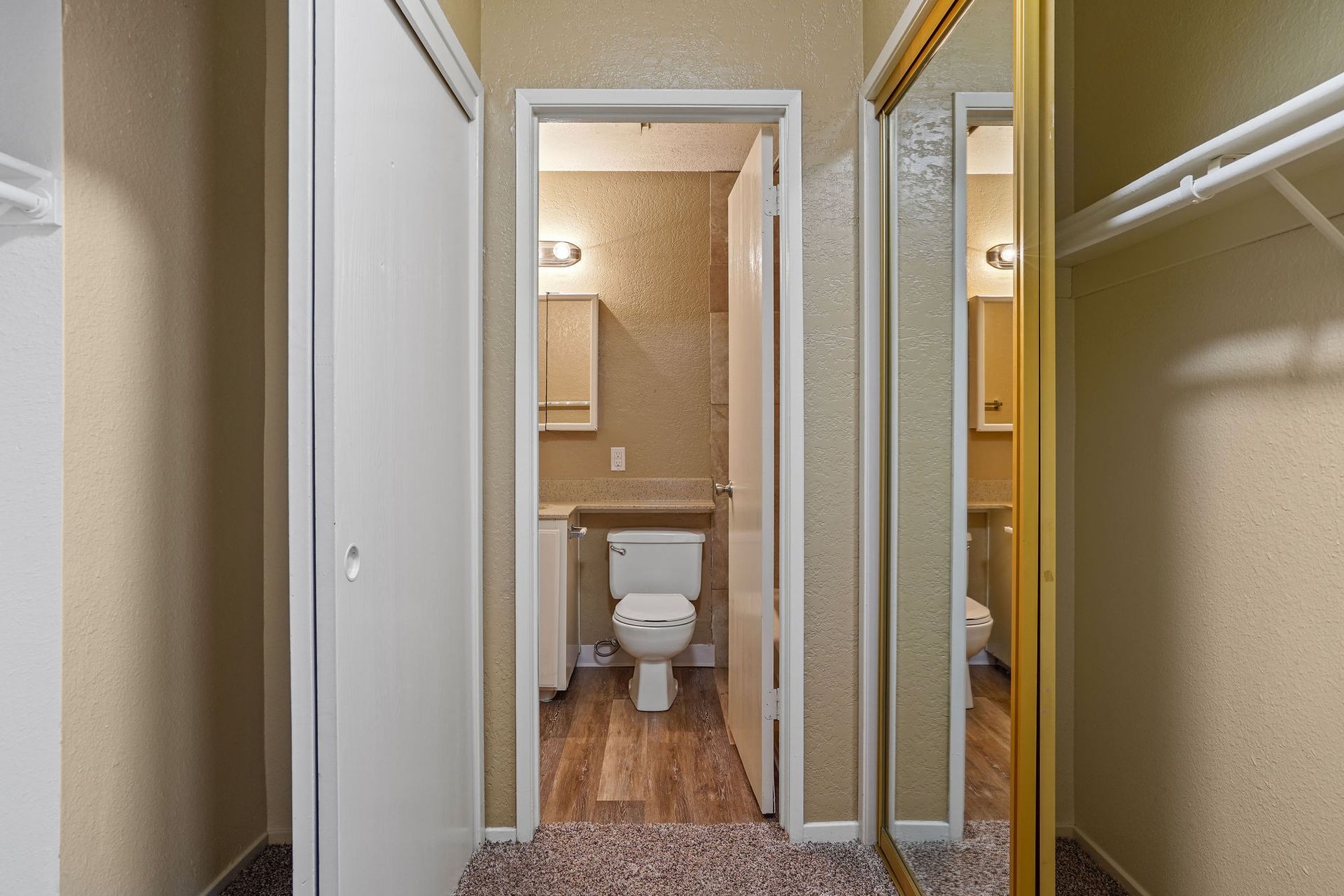 Narrow hallway view into a small bathroom with beige walls, toilet, wood floor, and mirrored closet doors.