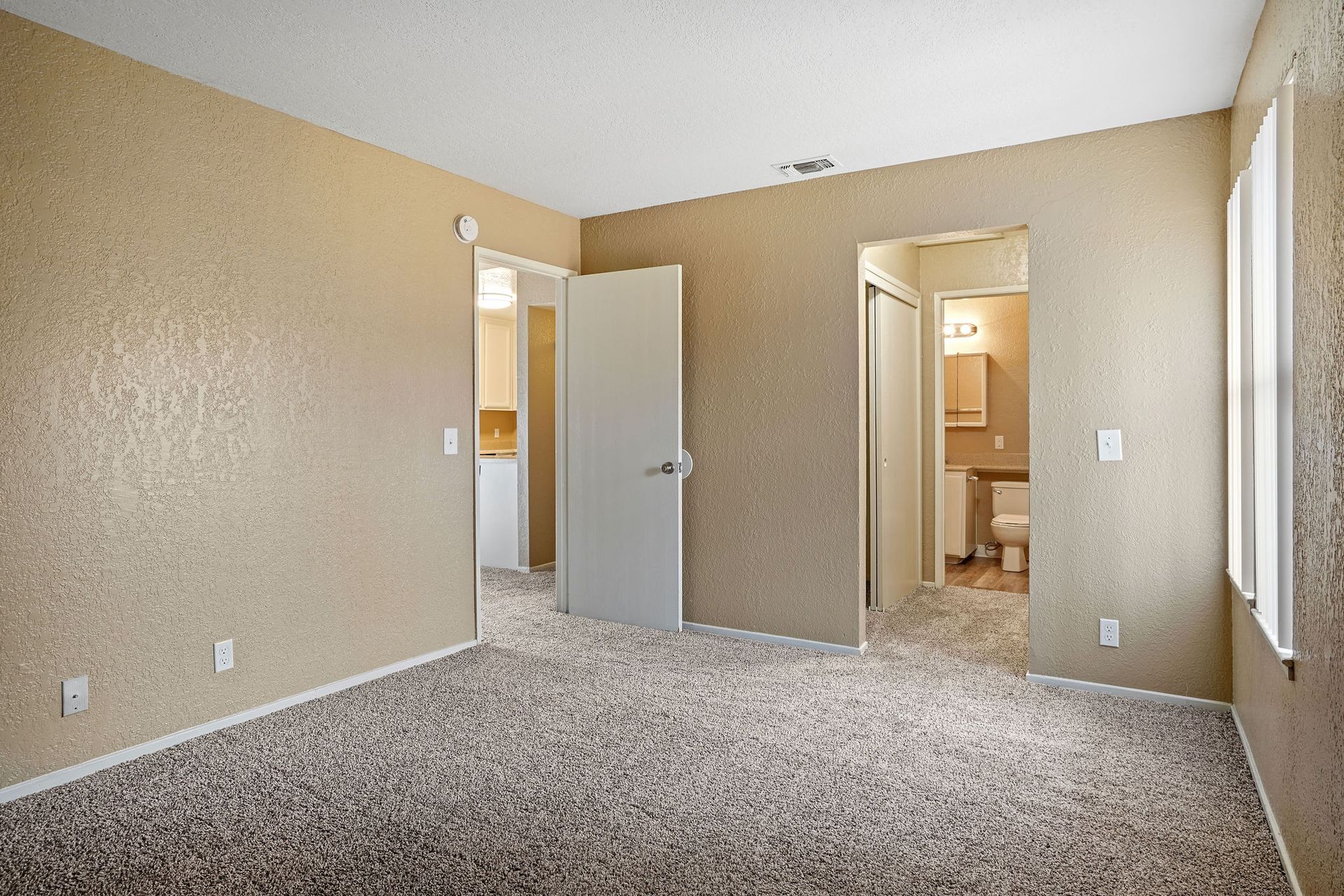 Empty beige bedroom with carpet, open doors, and a bathroom visible through the hallway.