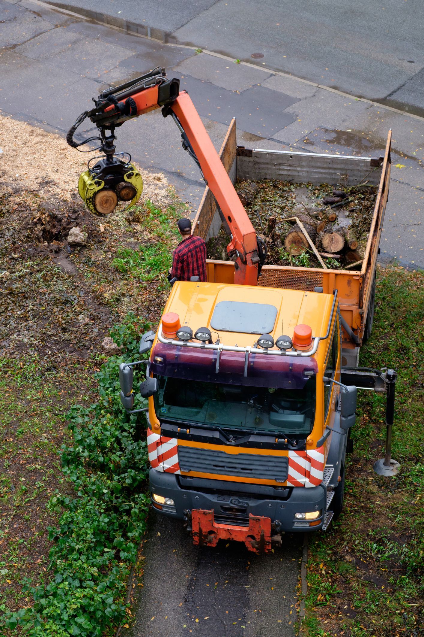 Yellow truck with an arm holding a cutting tool, loading debris into its bed, on a paved surface.