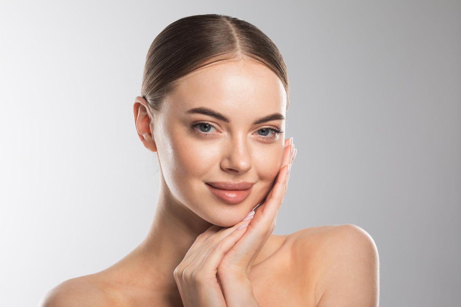 Woman with light skin, hands near face, smiling slightly. Against a grey backdrop.