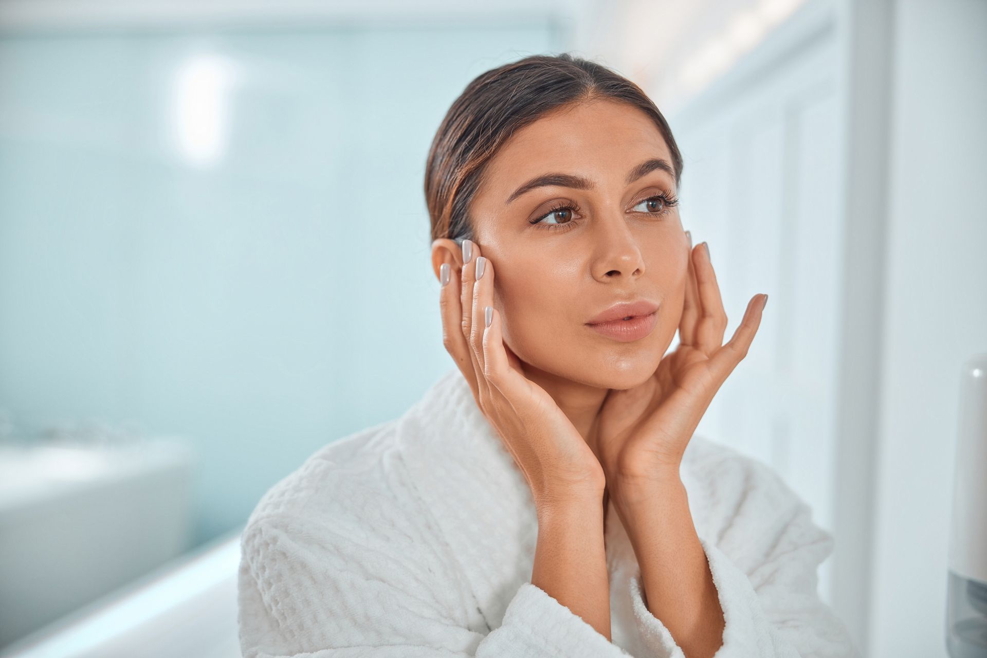 A woman in a bathrobe is looking at her face in the mirror.