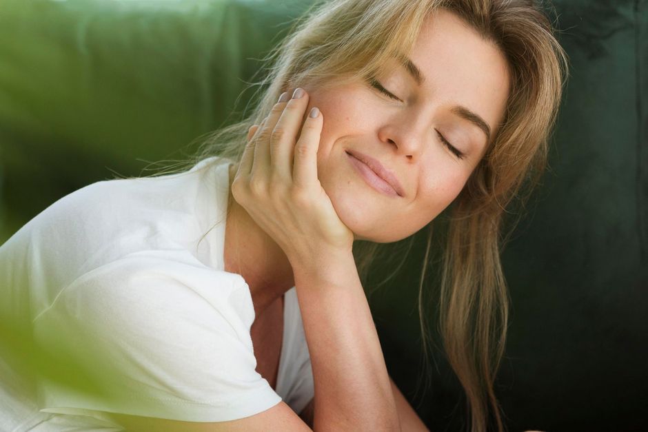 A woman is sitting on a couch with her eyes closed and her hand on her face.