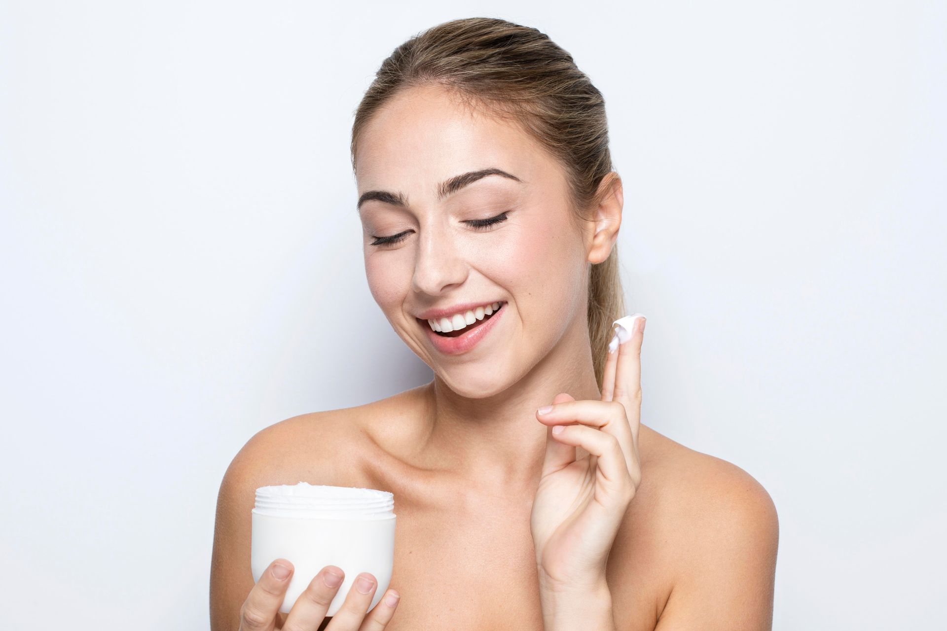 A woman is applying cream to her face while holding a jar.