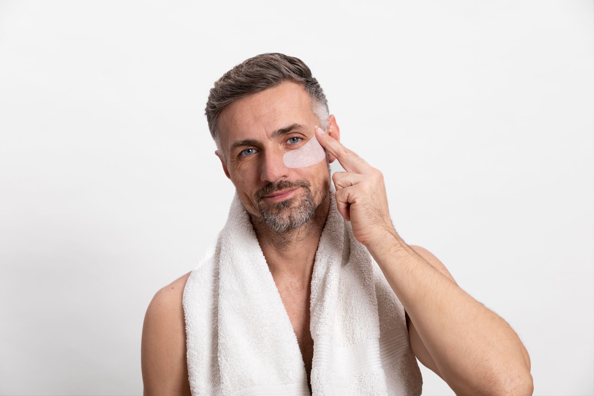 A man with a towel around his neck is applying eye patches to his face.