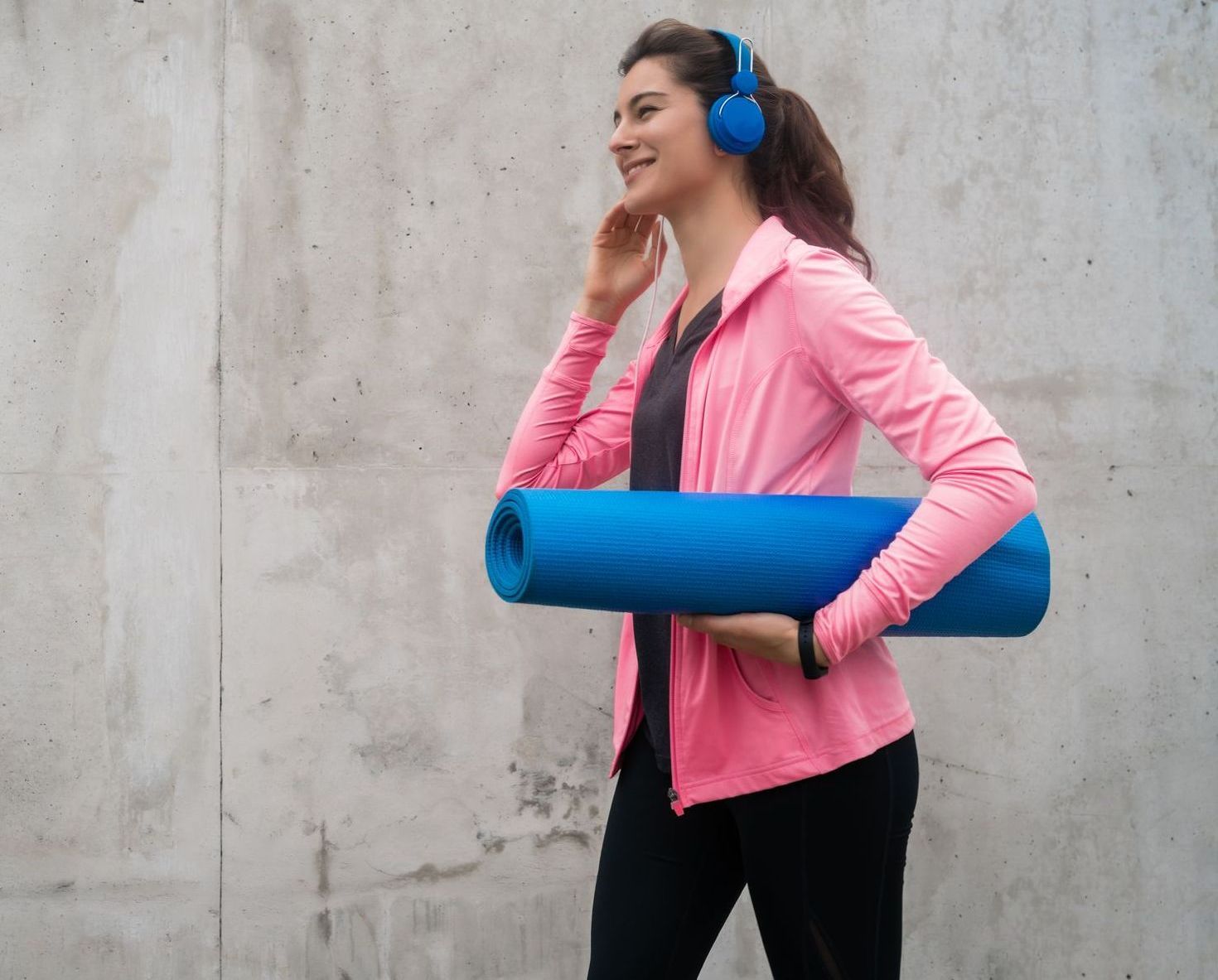 A woman wearing headphones is carrying a yoga mat and listening to music.