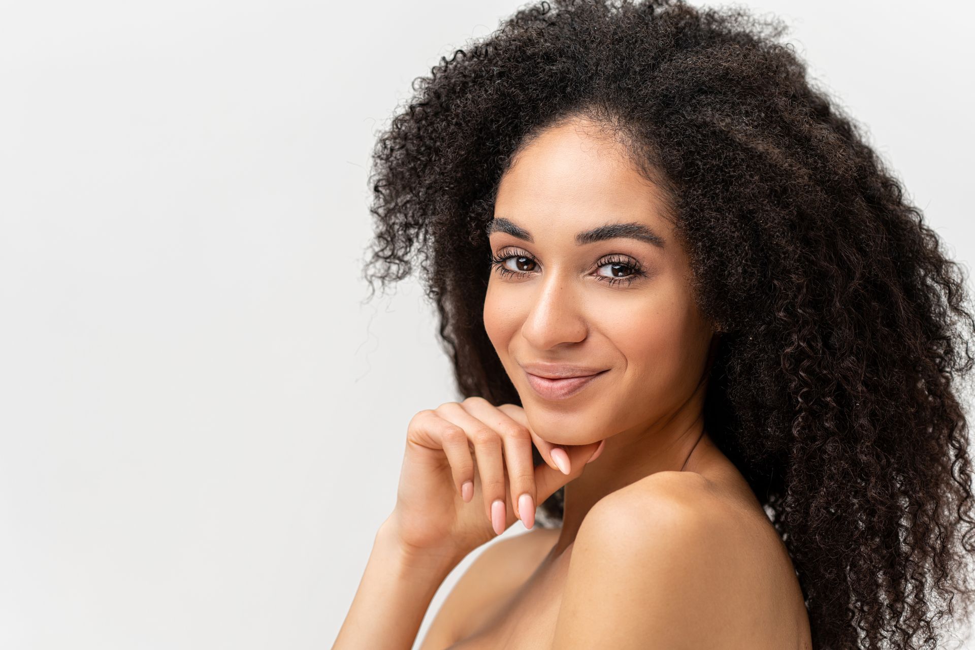 Woman with long, curly dark hair, smiles, and rests hand on chin, set against a white backdrop.