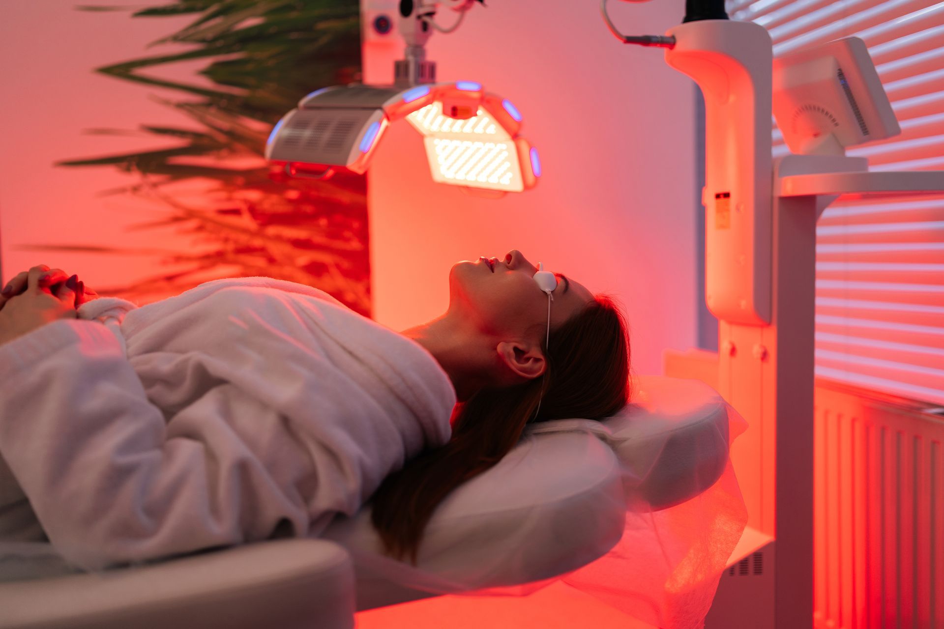 A woman is laying on a bed getting a red light treatment on her face.