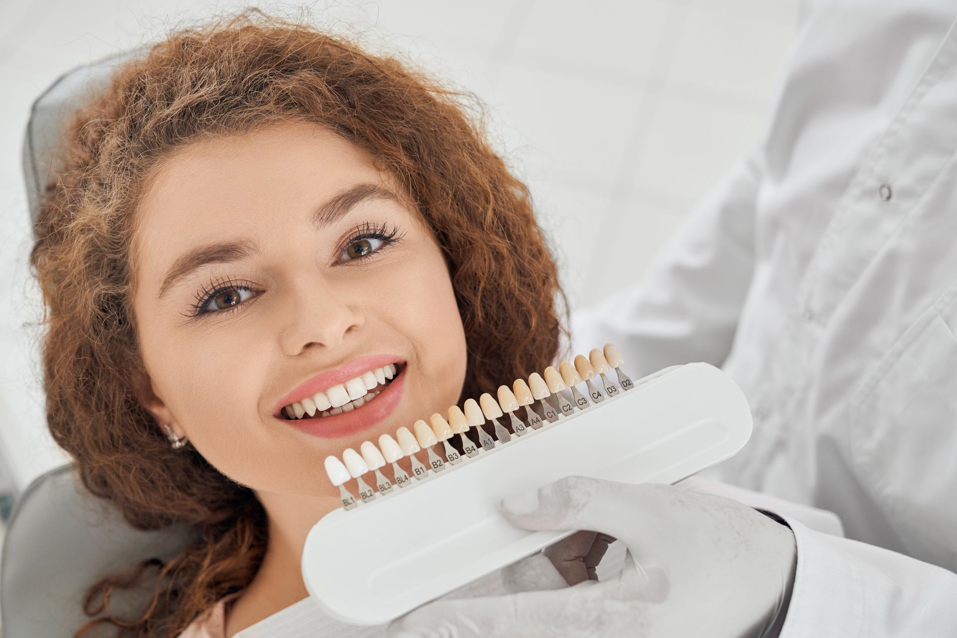 A woman is sitting in a dental chair holding a tooth color chart.