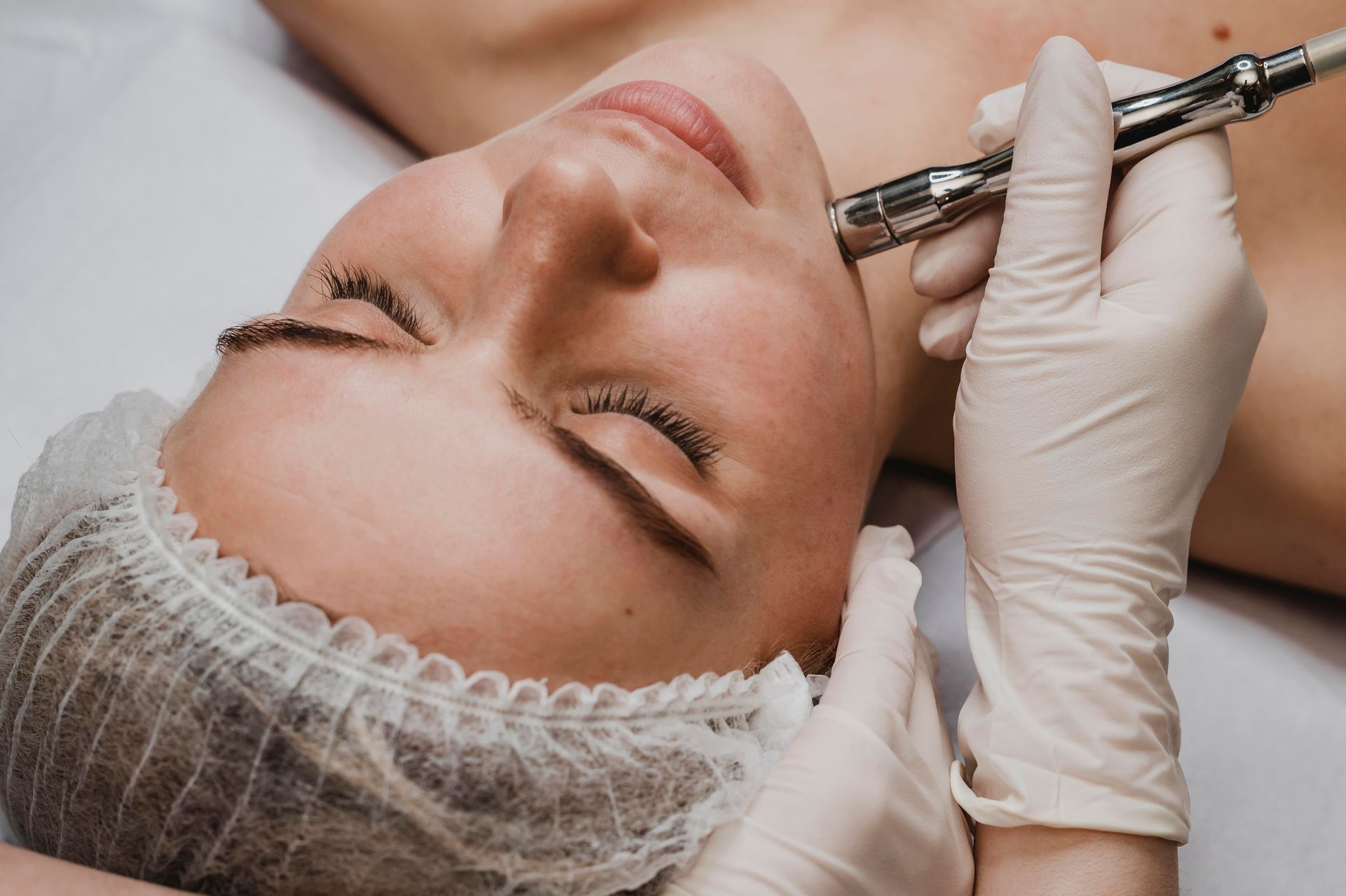 Woman receiving facial treatment; close-up shows device on her cheek. Person wearing gloves; medical setting.