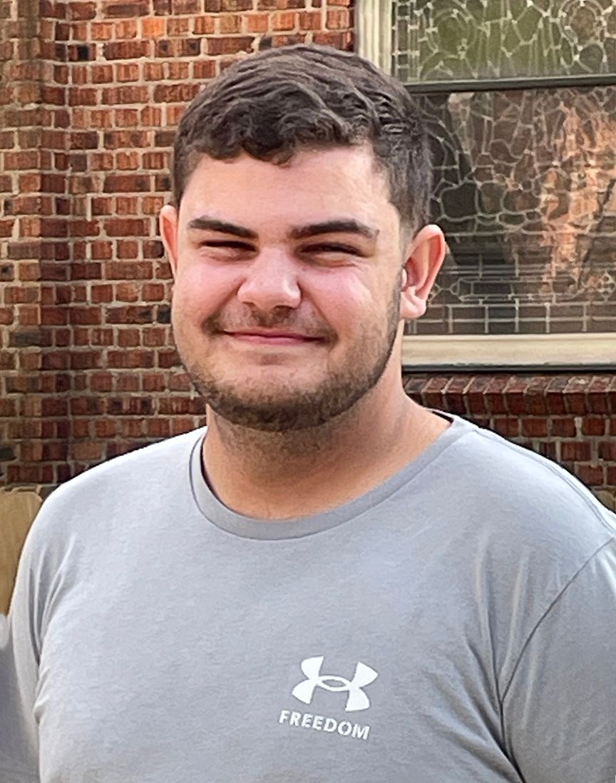 A man with a beard and a hat is standing in front of a brick building.