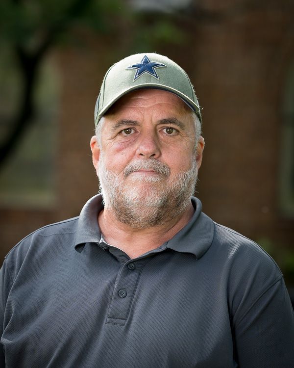 A man with a beard and a hat is standing in front of a brick building.