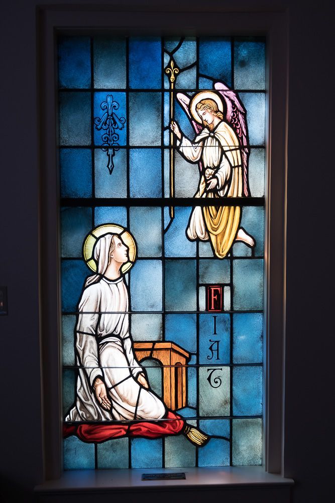 A stained glass window shows a woman kneeling in front of an angel holding a cross.