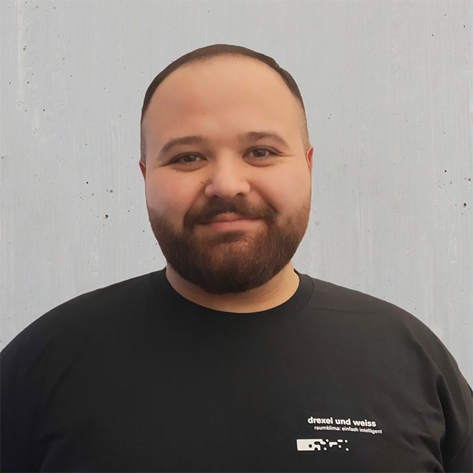 Ömer Mermer steht vor einer hellen Wand, trägt ein schwarzes T-Shirt mit Logo, hat sehr kurze Haare und einen akkurat geschnittenen Bart. Er lächelt sehr freundlich in die Kamera.