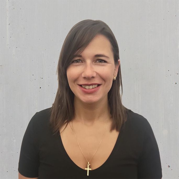 Ein Mann in schwarzem T-Shirt lächelt in die Kamera, er steht vor einer grauen Wand.