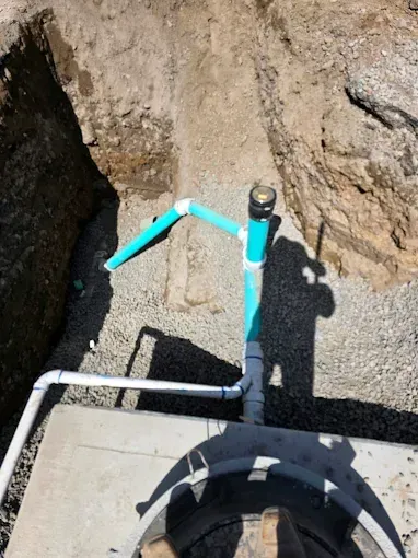 A high-angle view of a construction trench showing PVC pipes connected to a concrete manhole structure.
