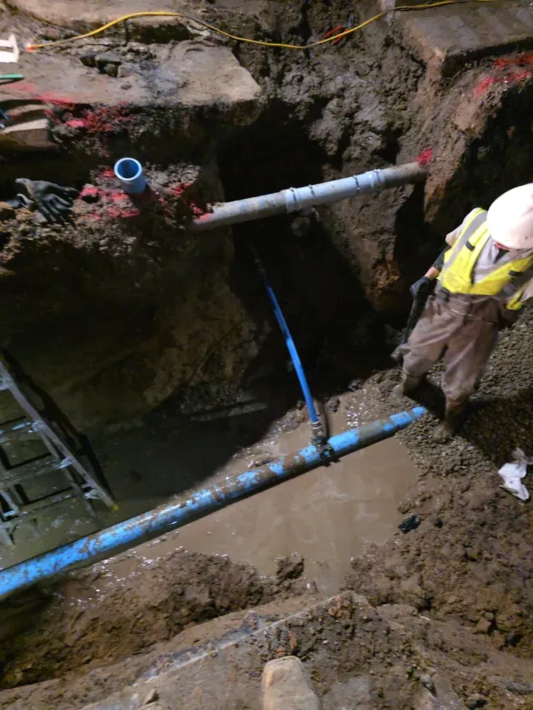 A worker in a high-visibility vest and hard hat stands beside a construction trench containing blue water pipes.