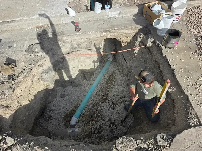A person in a trench uses a level to check a pipe installation, with construction supplies visible nearby.