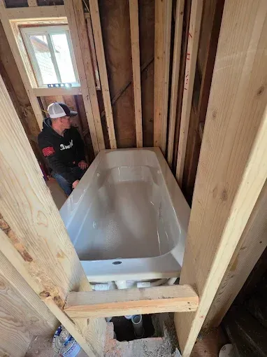 A person wearing a hooded sweatshirt kneels in a wooden framed bathroom alcove, installing a new white bathtub.