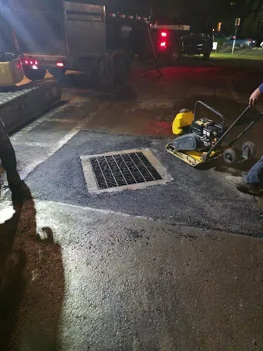 A worker uses a yellow plate compactor to smooth fresh asphalt around a storm drain at night.