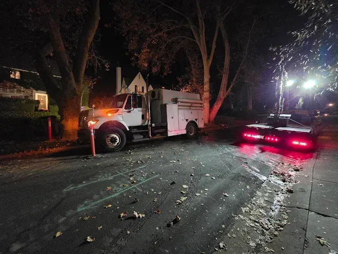 A white utility truck is parked on a residential street at night, with a trailer attached that has red taillights glowing.
