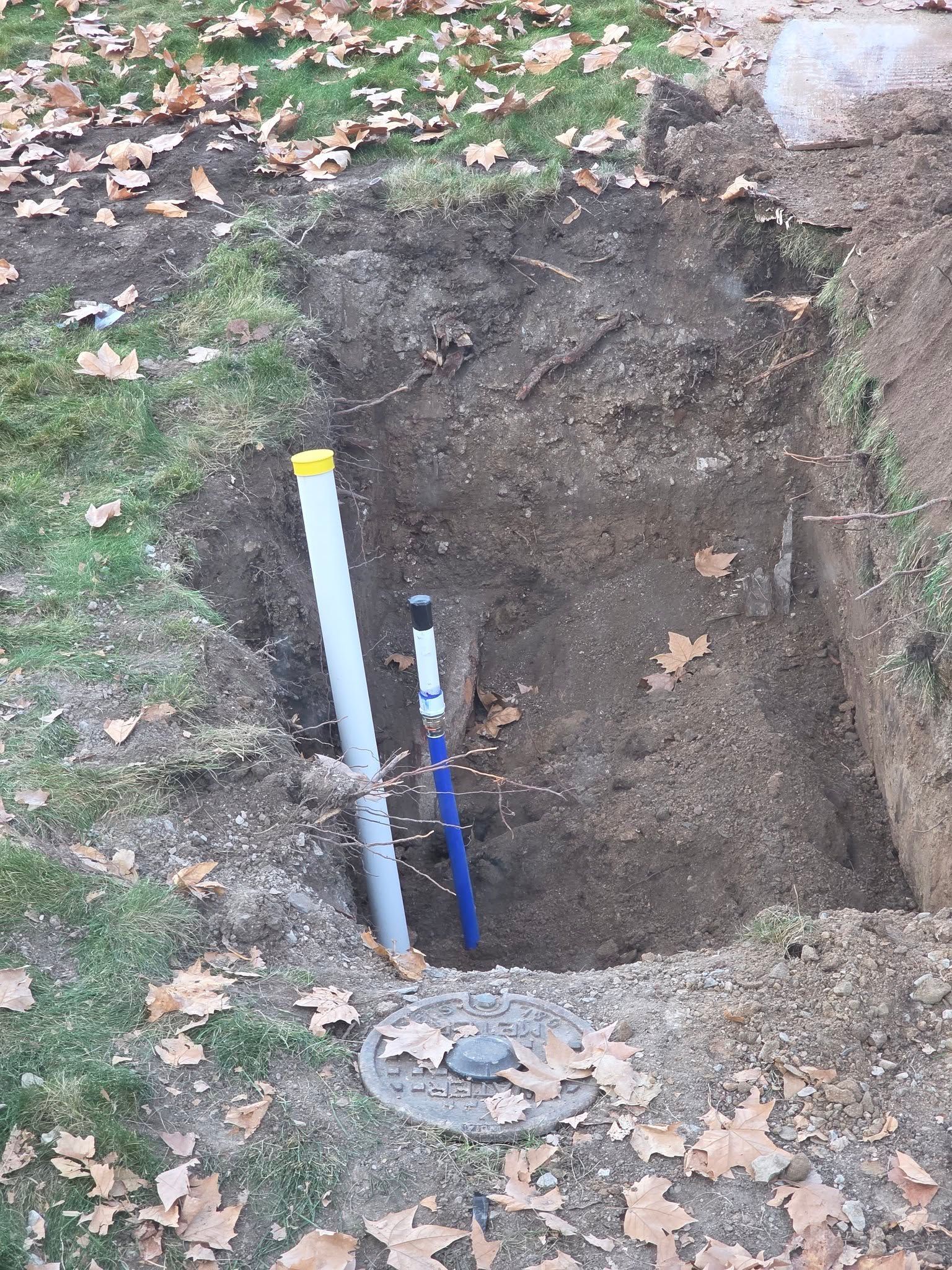 A hole in the ground reveals a gray pipe with a yellow cap and a blue-handled shutoff valve next to a concrete cover.
