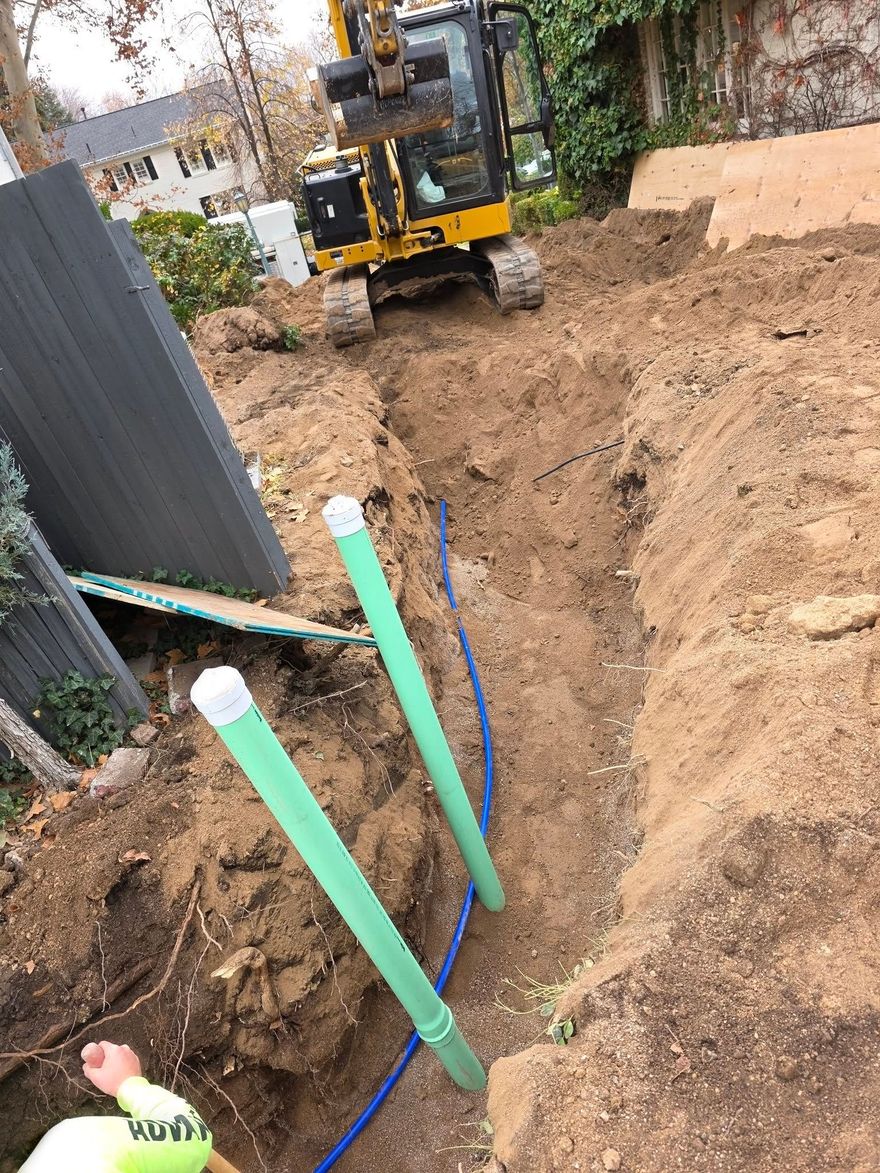 A yellow excavator sits at the end of a dirt trench where green pipes and a blue line are being installed in a backyard.