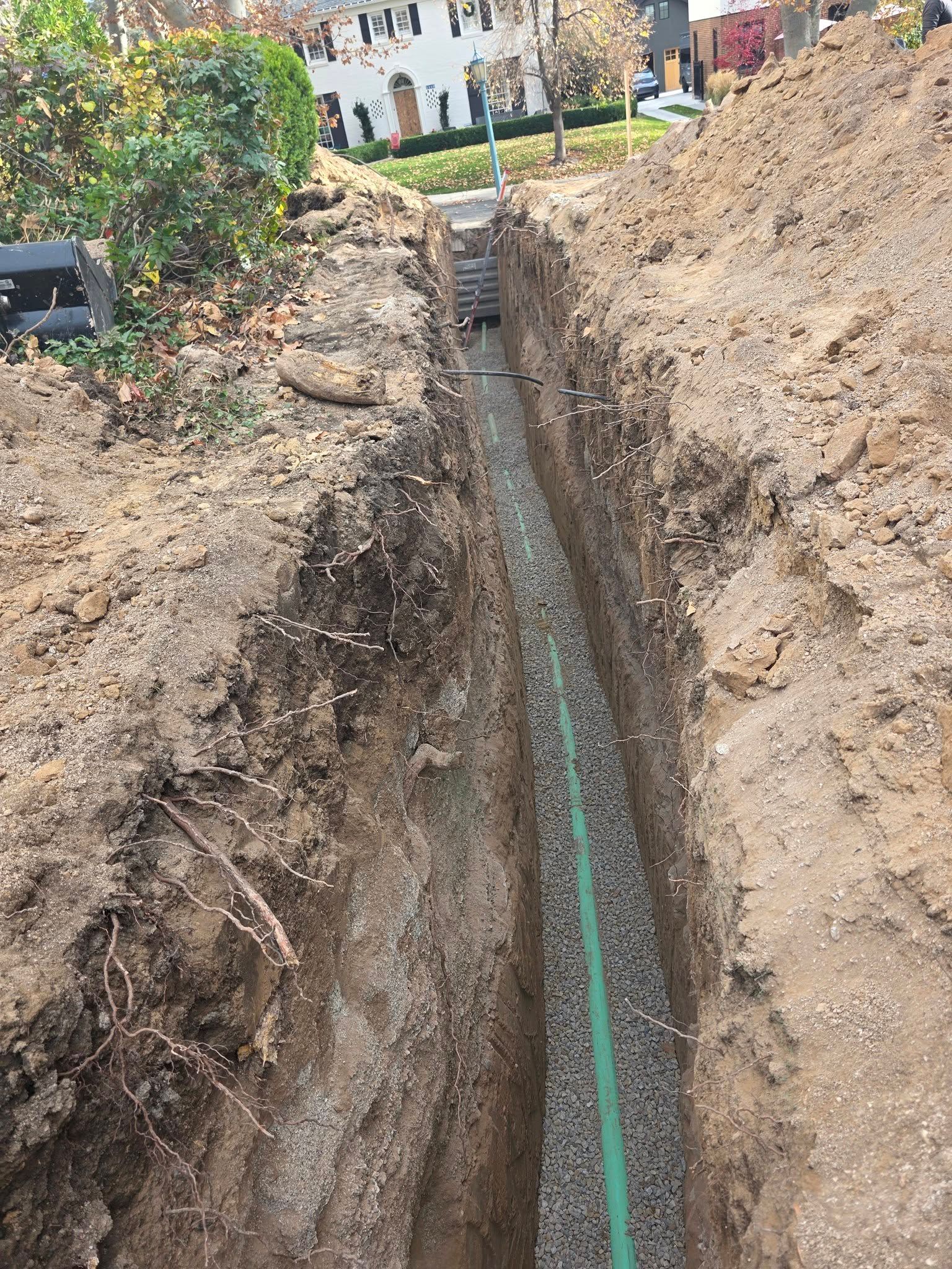 A narrow trench in the ground containing a green pipe and loose gravel, with dirt piles on either side.