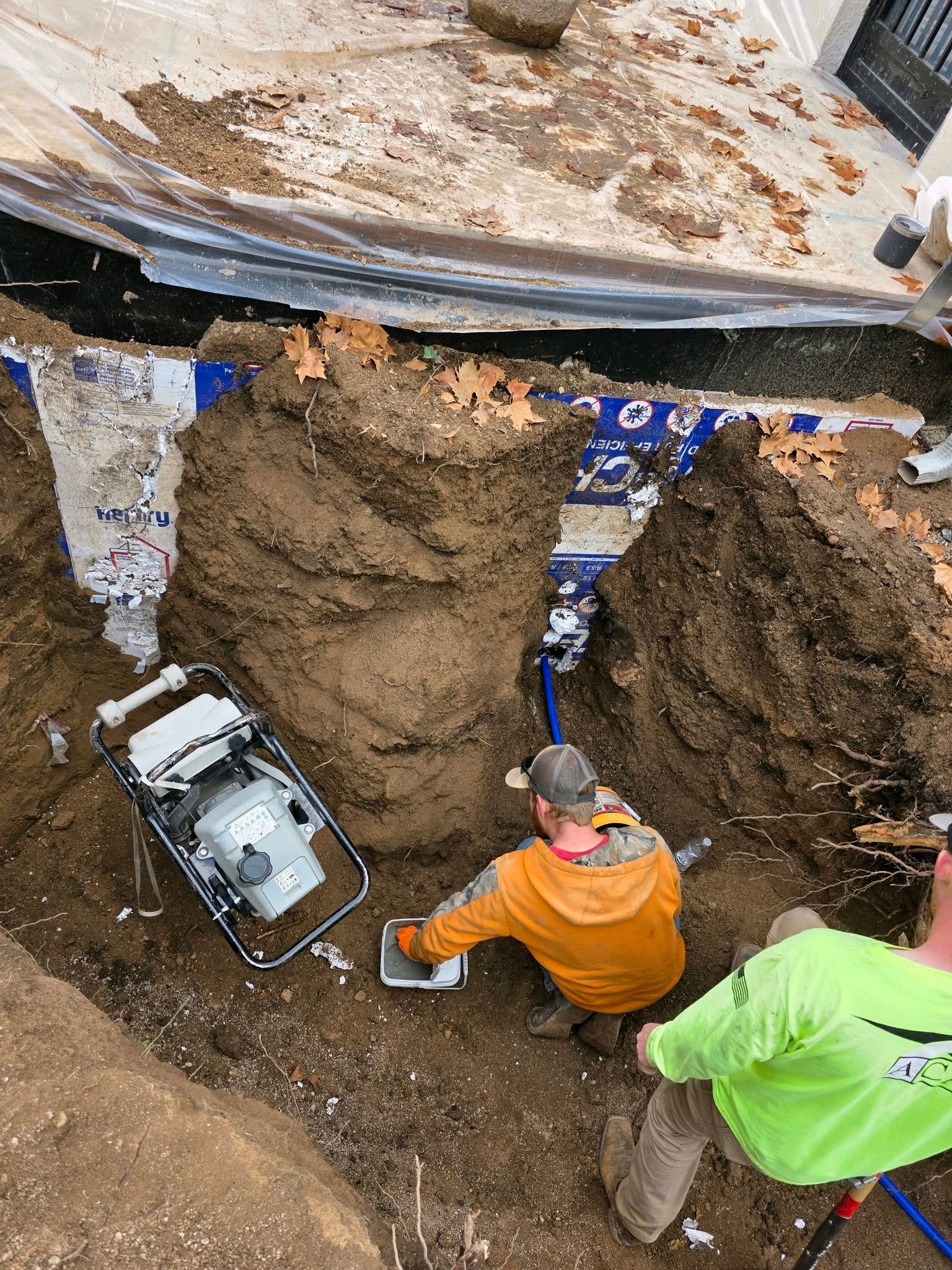 Two workers in a construction trench, one operating a piece of machinery, next to exposed foundation walls.