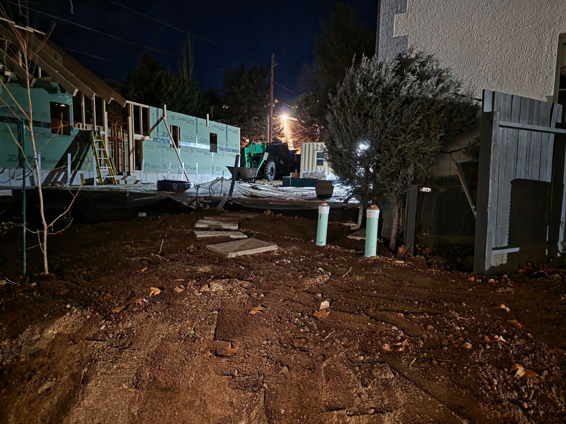 A night view of a muddy yard with a partially constructed house, green insulation panels, and two white pipes in the soil.