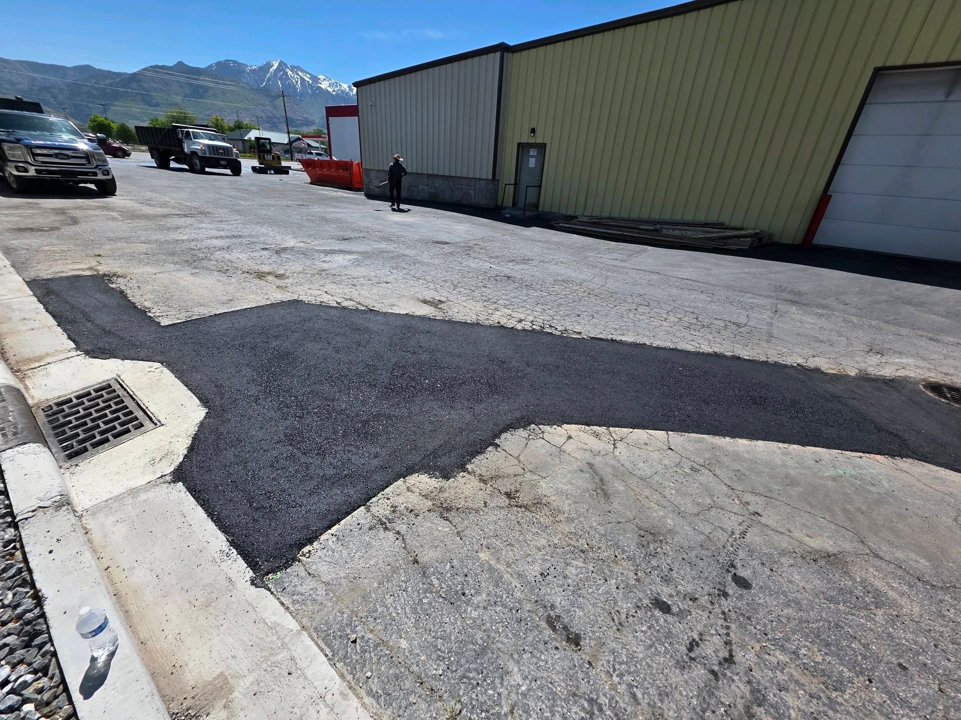 A patch of fresh dark asphalt on a worn, light-colored gravel lot near an industrial building with mountains beyond.