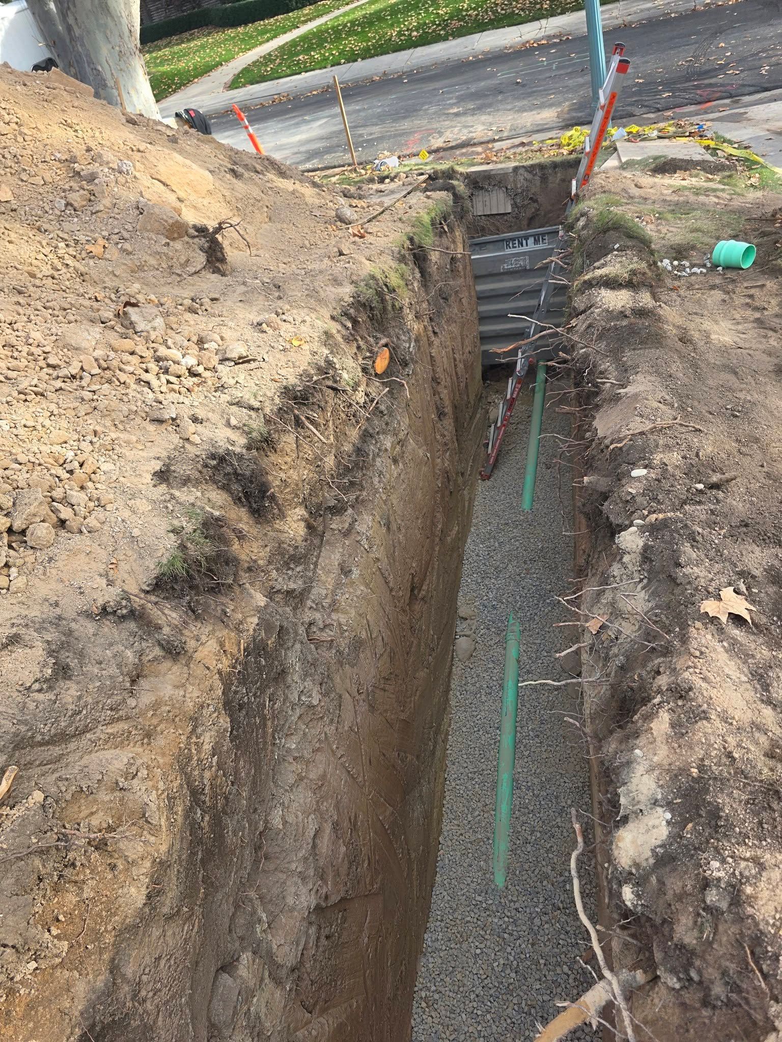 A narrow, deep trench cut into soil, featuring a gravel base and a green pipe running along the bottom.