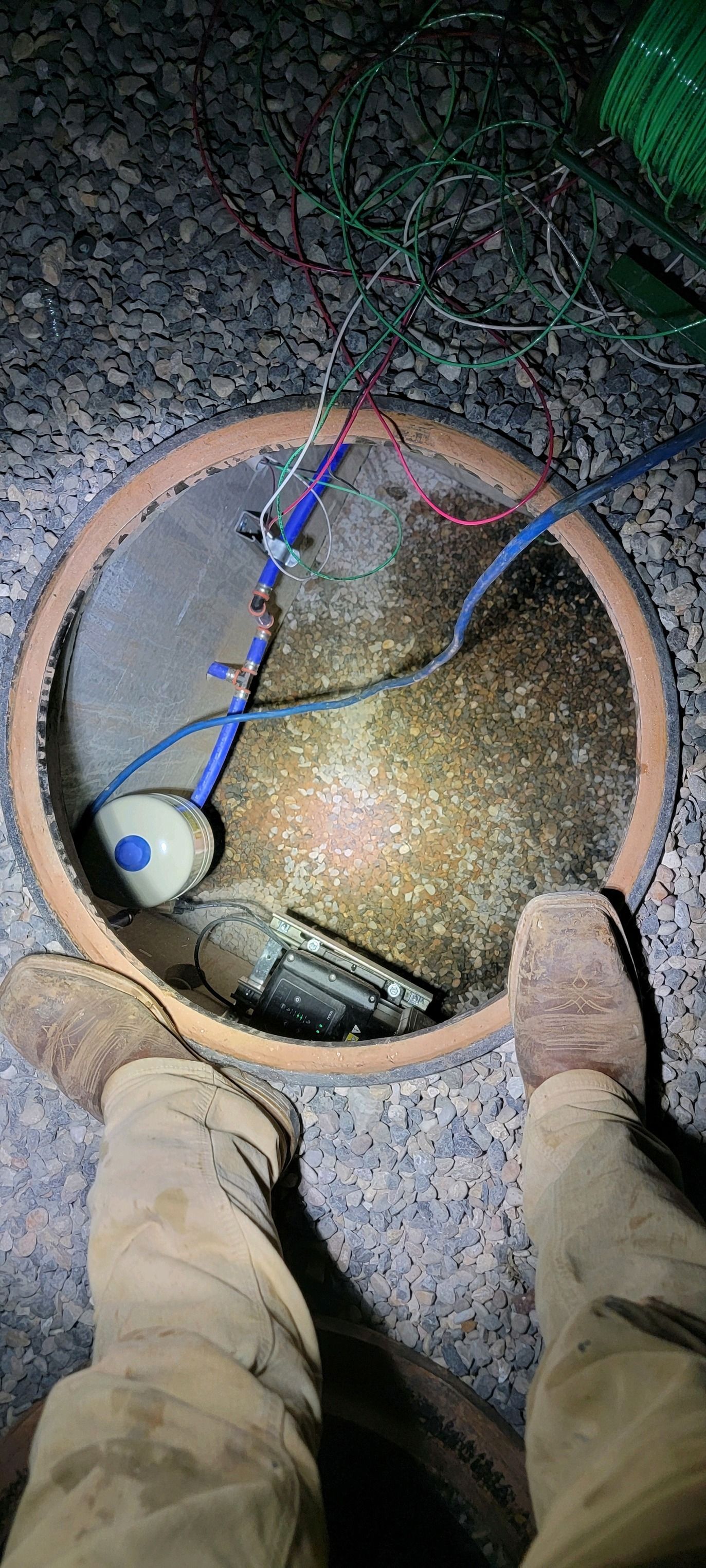A person’s legs stand over an open, circular in-ground utility box containing wires and a float switch.