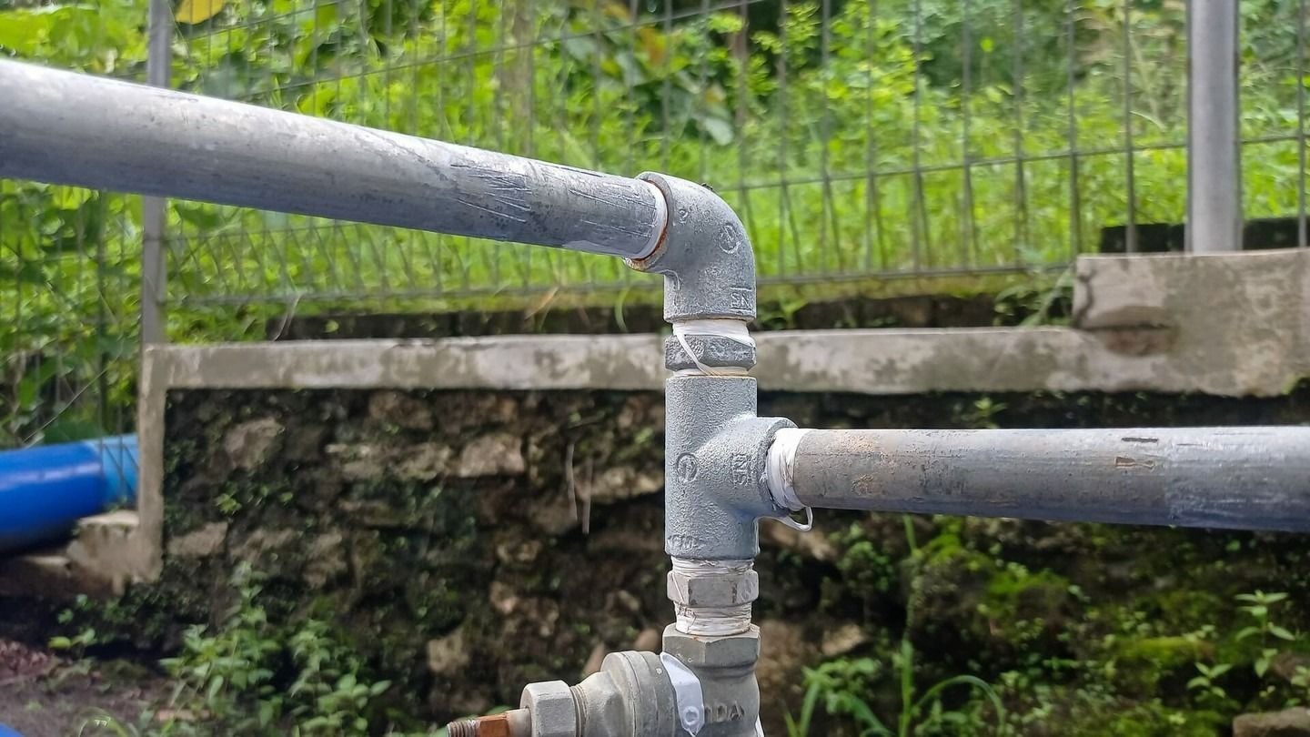 A close-up of gray metal plumbing pipes with a T-junction and elbow fitting, covered in water droplets outdoors.