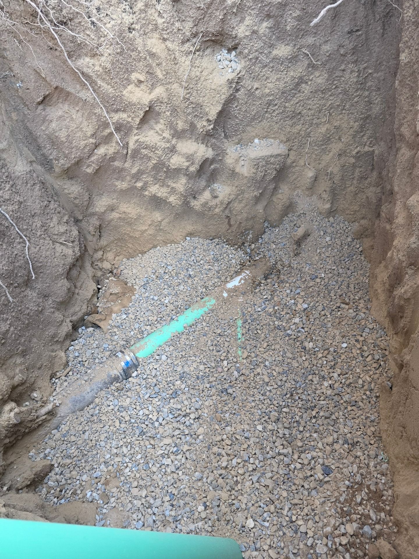 A green plastic pipe segment connected to a larger pipe, resting on a bed of gravel inside a deep, open earthen trench.