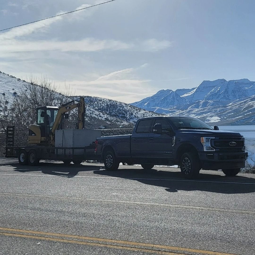 A dark pickup truck tows a flatbed trailer with a small yellow excavator past a snowy mountain range and water.