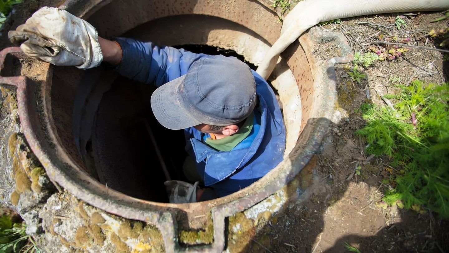 A worker in a blue uniform and cap reaches into an open manhole while a white hose runs into the opening.