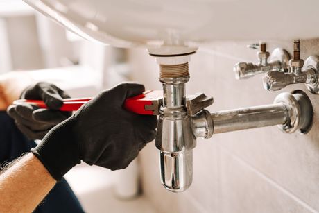A plumber wearing black work gloves uses a red wrench to tighten a metal pipe under a sink.