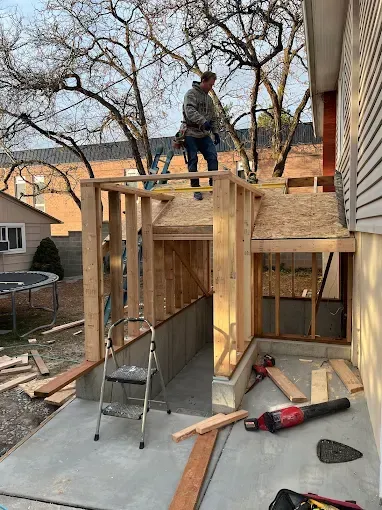 A worker stands on the wooden frame of a home addition project in a backyard, near a ladder and construction materials.