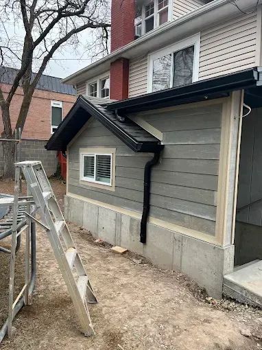 An exterior view of a house addition with gray horizontal siding, a white-framed window, and a black-trimmed roof.