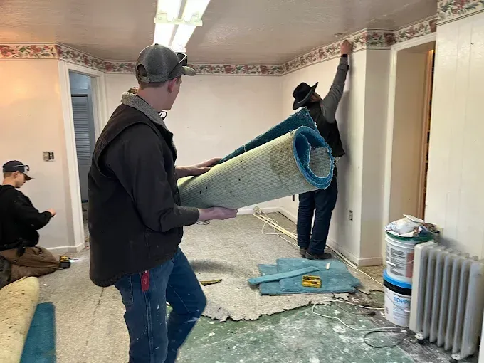 Three workers renovate a room, removing old carpet and wallpaper, with rolls of flooring and supplies on the floor.