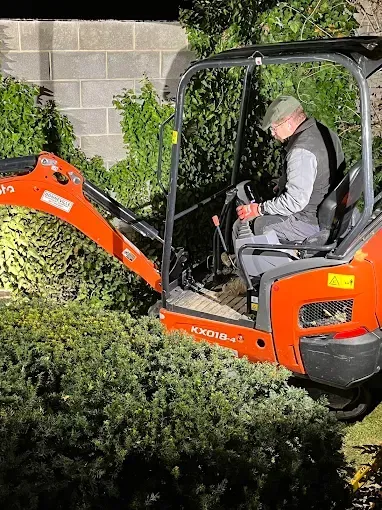A person operating an orange Kubota excavator in a residential backyard near a concrete block wall and greenery.
