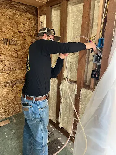 A worker in a black long-sleeve shirt installs electrical wiring into wall boxes in a room with spray foam insulation.