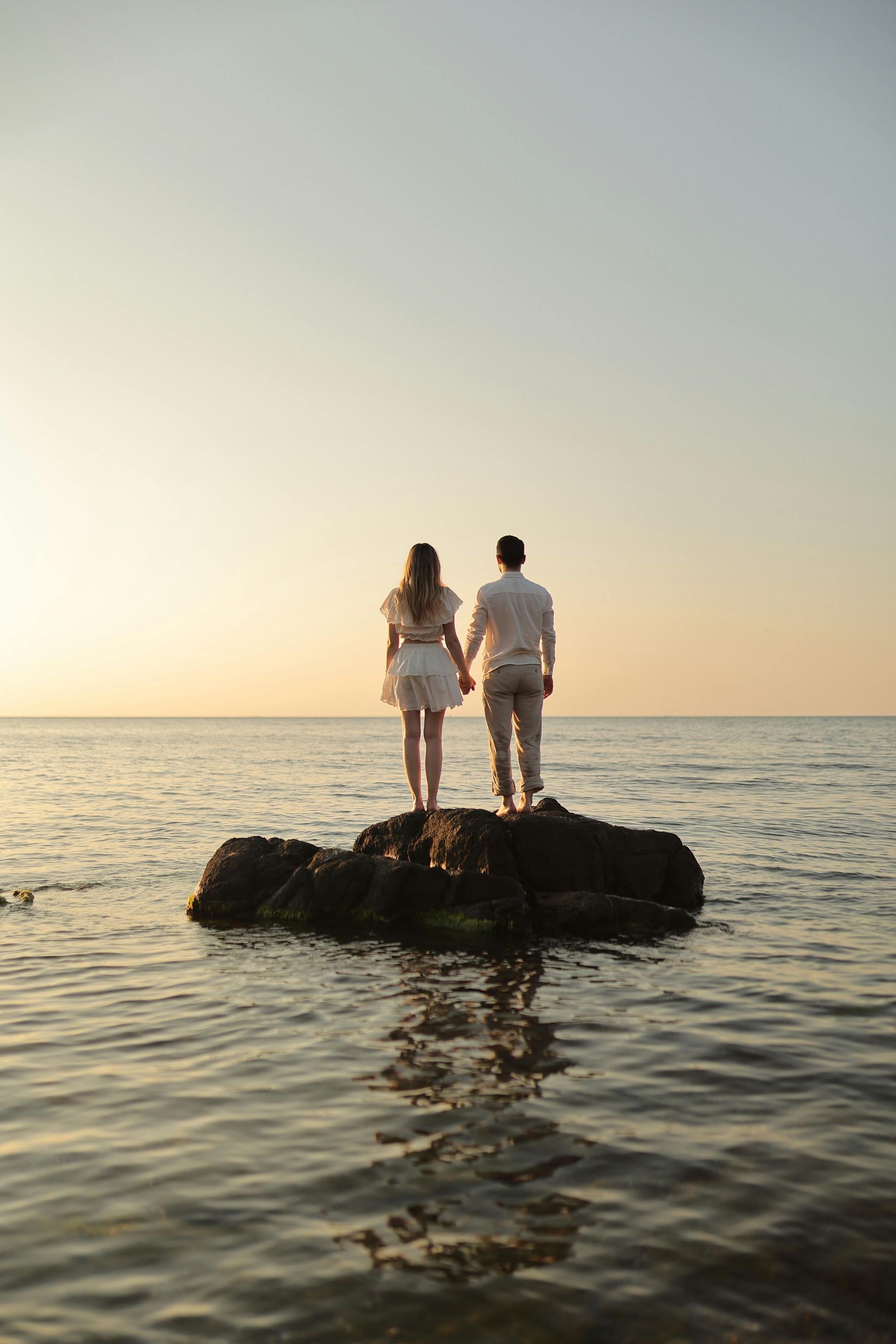 A man and a woman are standing on a rock in the middle of the ocean holding hands.