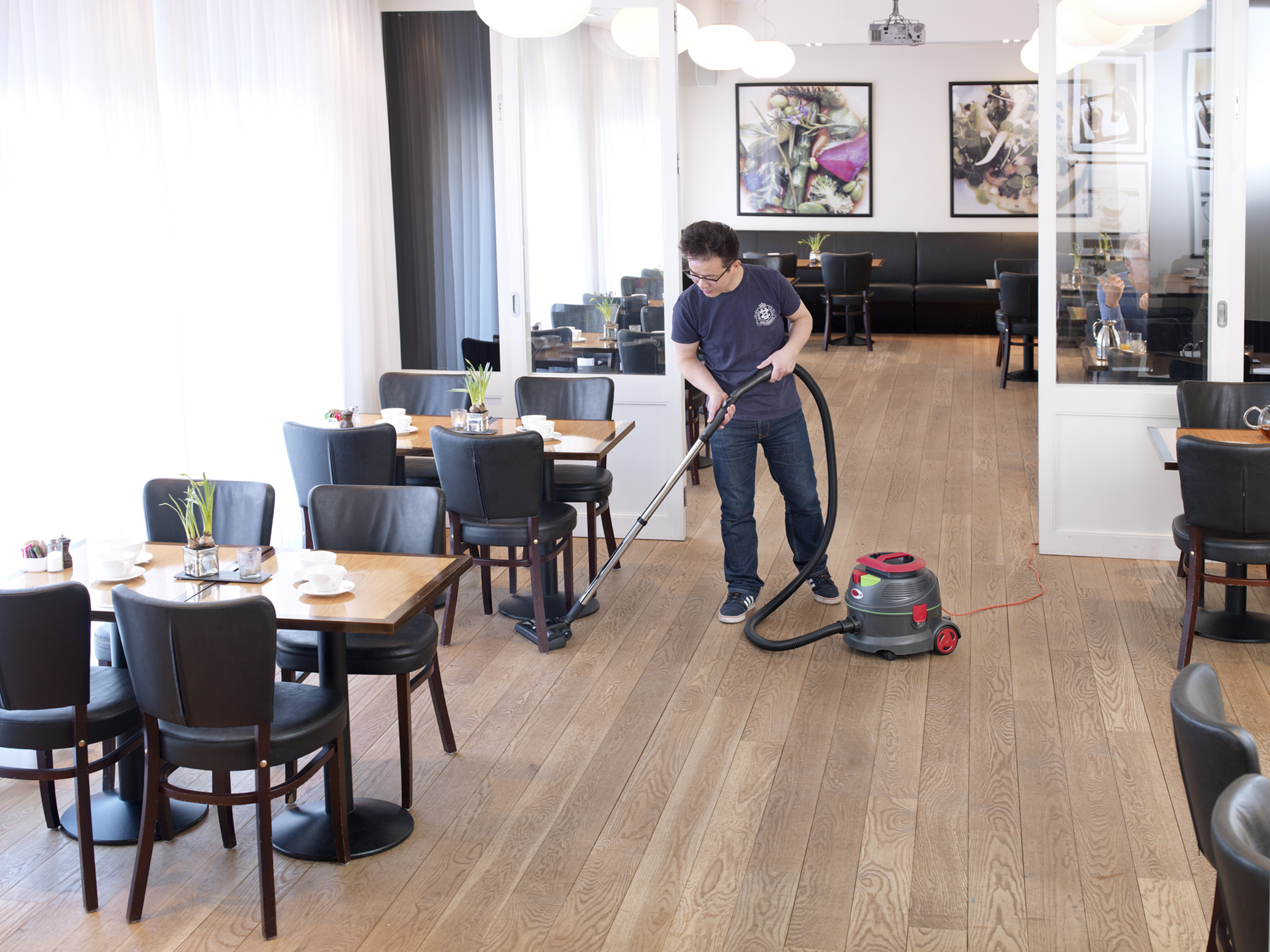 A person vacuums a restaurant floor between tables. The room is brightly lit, with wooden floors.