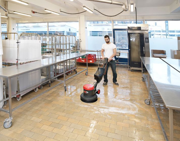 Man using floor cleaning machine in a commercial kitchen with stainless steel tables and equipment.