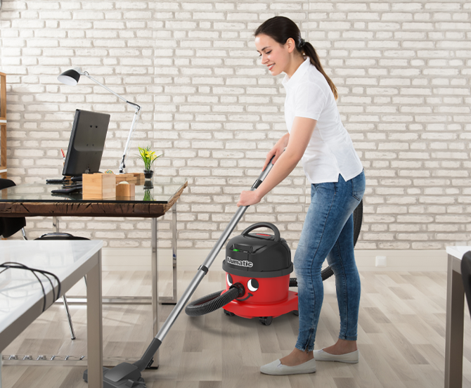 Woman vacuuming a wood floor in an office setting with red vacuum cleaner.